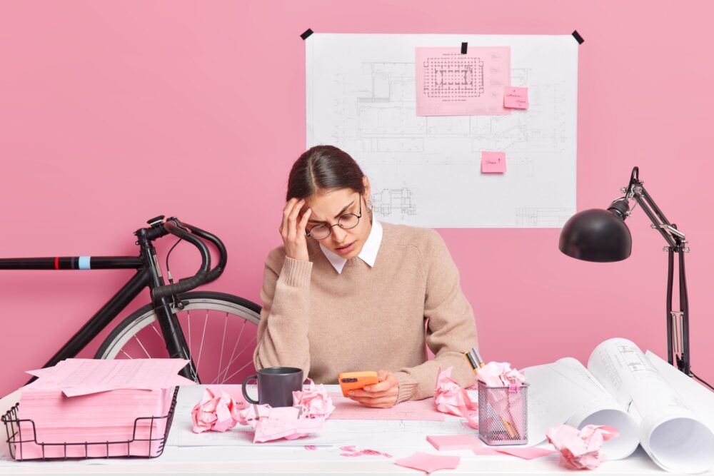 Overworked tired female architect focused at smartphone has much work to do works on architectural p